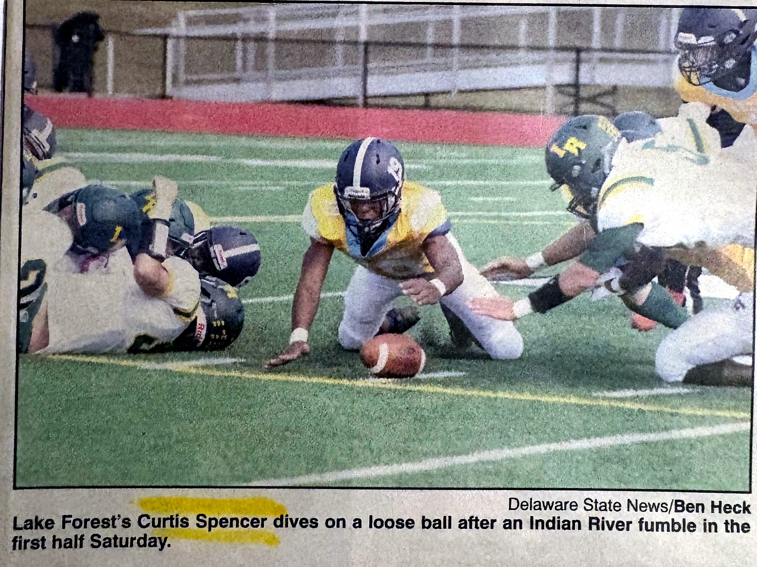 Football players dive for a loose ball on a green turf during a game; a central player in a yellow jersey and blue helmet reaches for the ball as teammates close in.