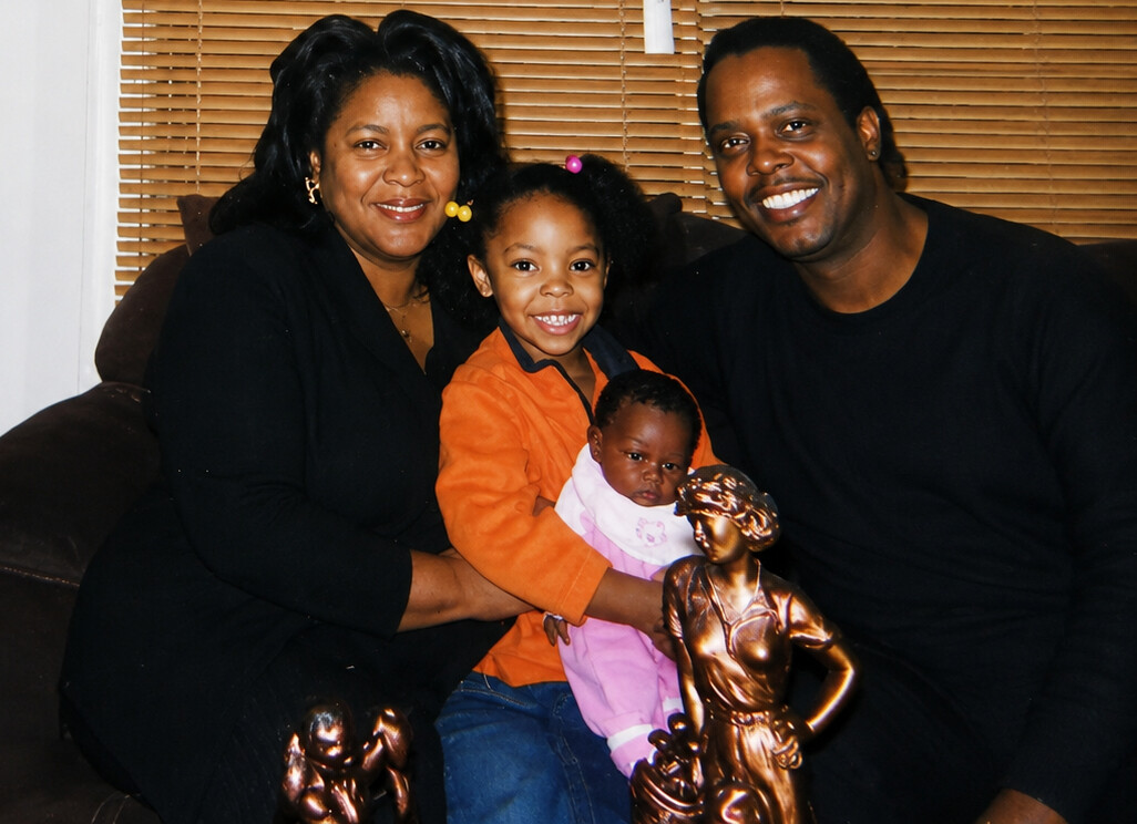 Smiling family of four on a couch: mother, father, an older daughter in orange and a baby girl in pink, with bronze statues in front.