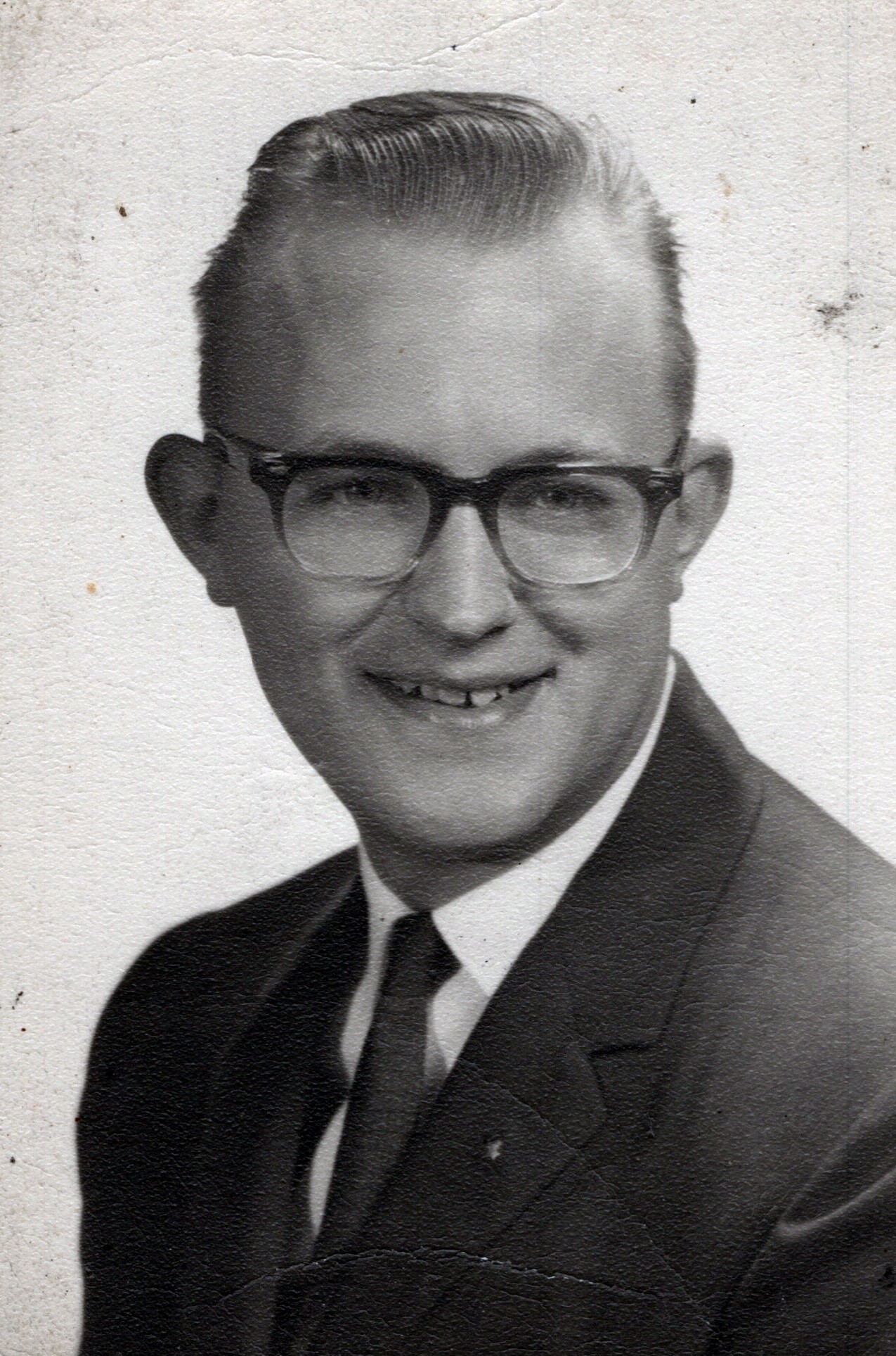 Young man in a suit and tie, wearing glasses, posed for a black-and-white portrait.