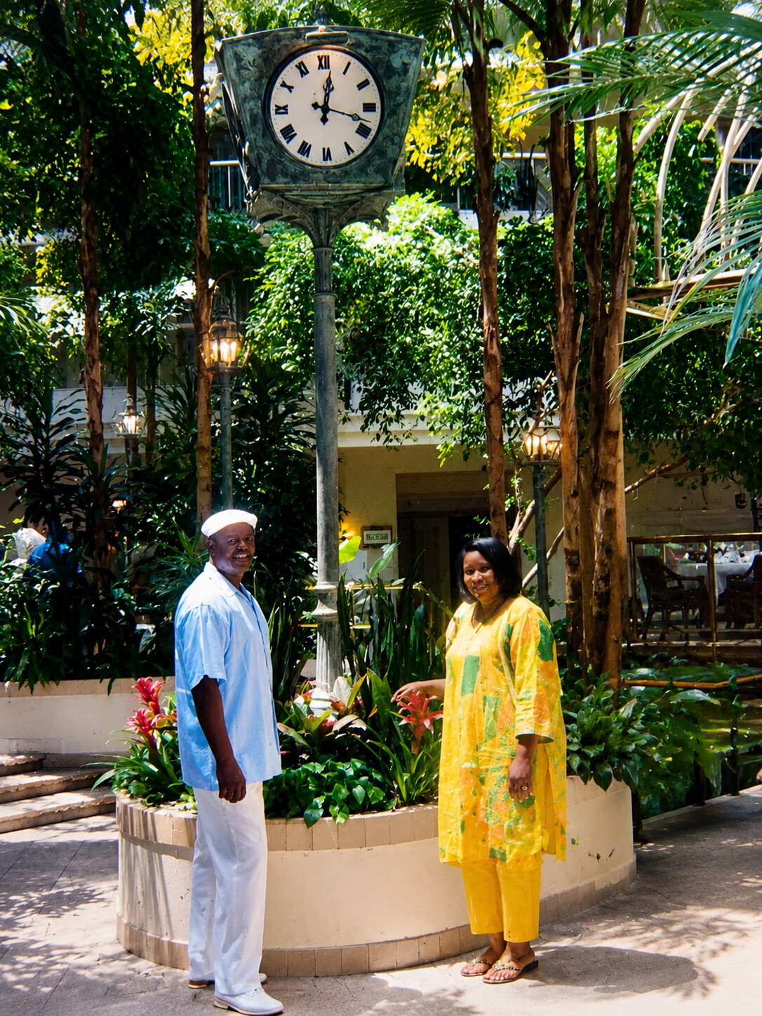 A man in a light blue shirt and white pants and a woman in a bright yellow outfit pose beside a circular planter in a tropical courtyard, with a large vintage clock on a pedestal behind them.