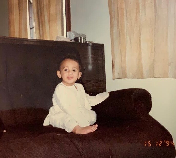 Baby sitting cross‑legged on a dark brown couch, wearing white pajamas and smiling in a cozy living room.