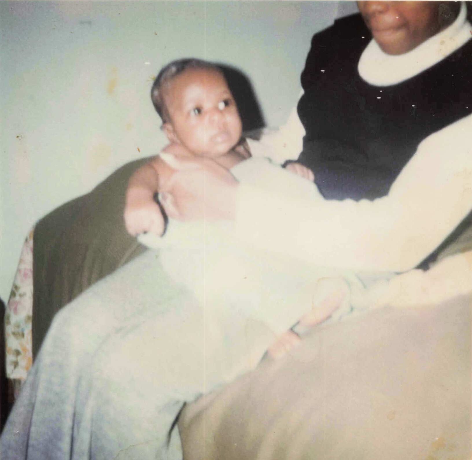 Infant cradled in the arms of an adult, facing left, in a pale room setting.