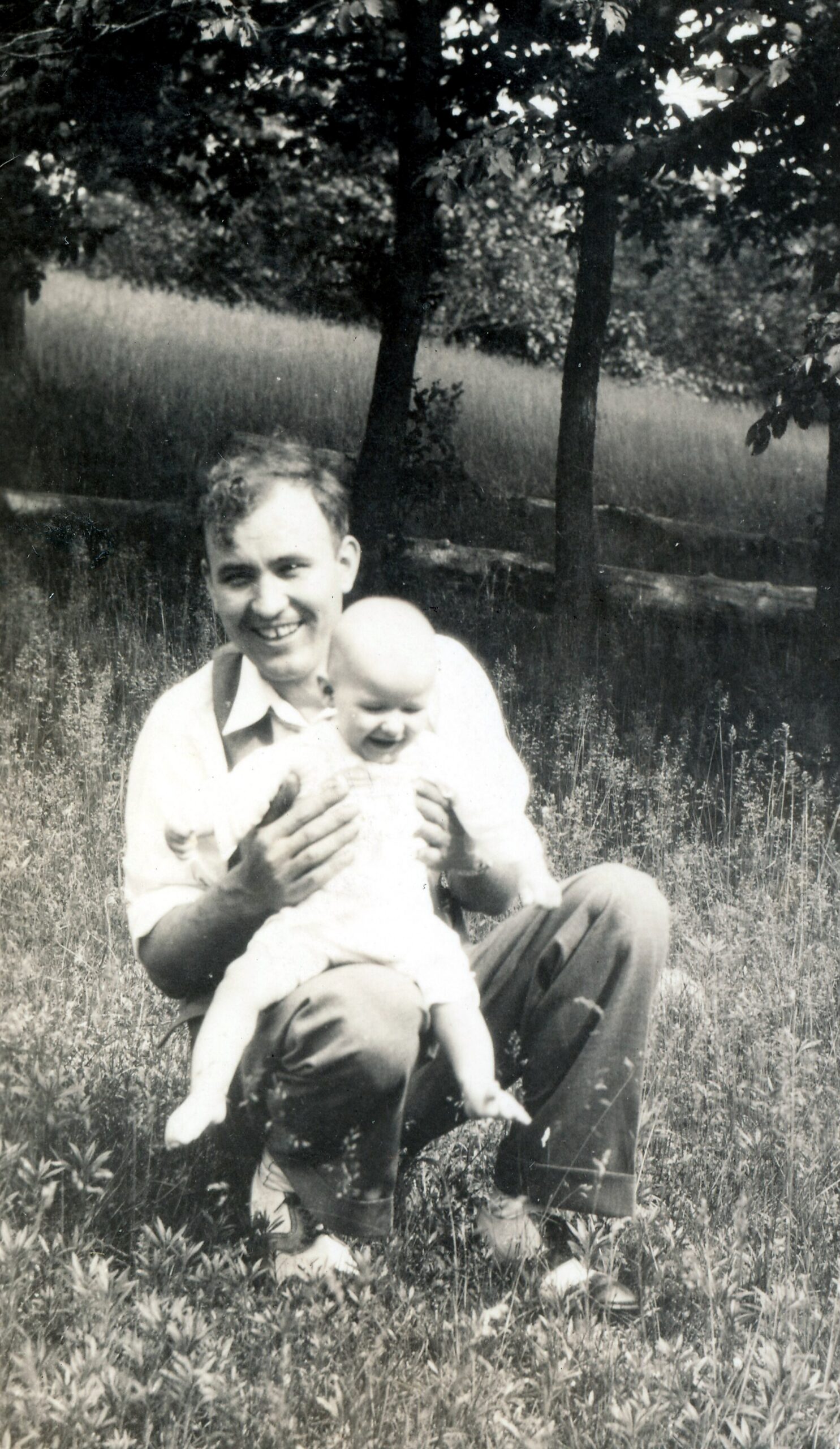 Man sitting in a grassy field outdoors, smiling while cradling a baby in his arms with trees in the background.