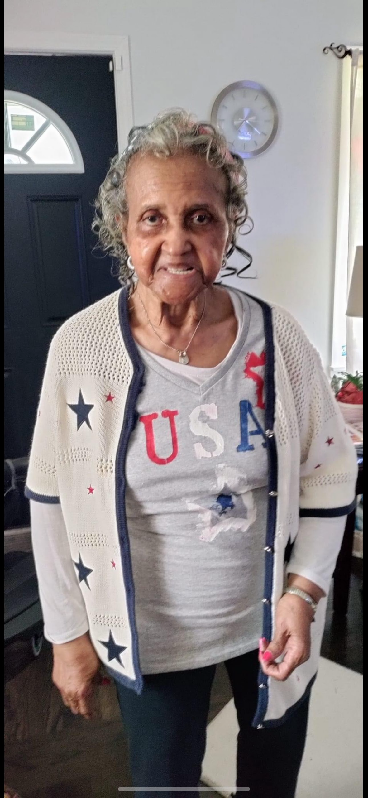 Older woman with gray hair standing indoors, wearing a white star-pattern cardigan over a gray USA shirt.