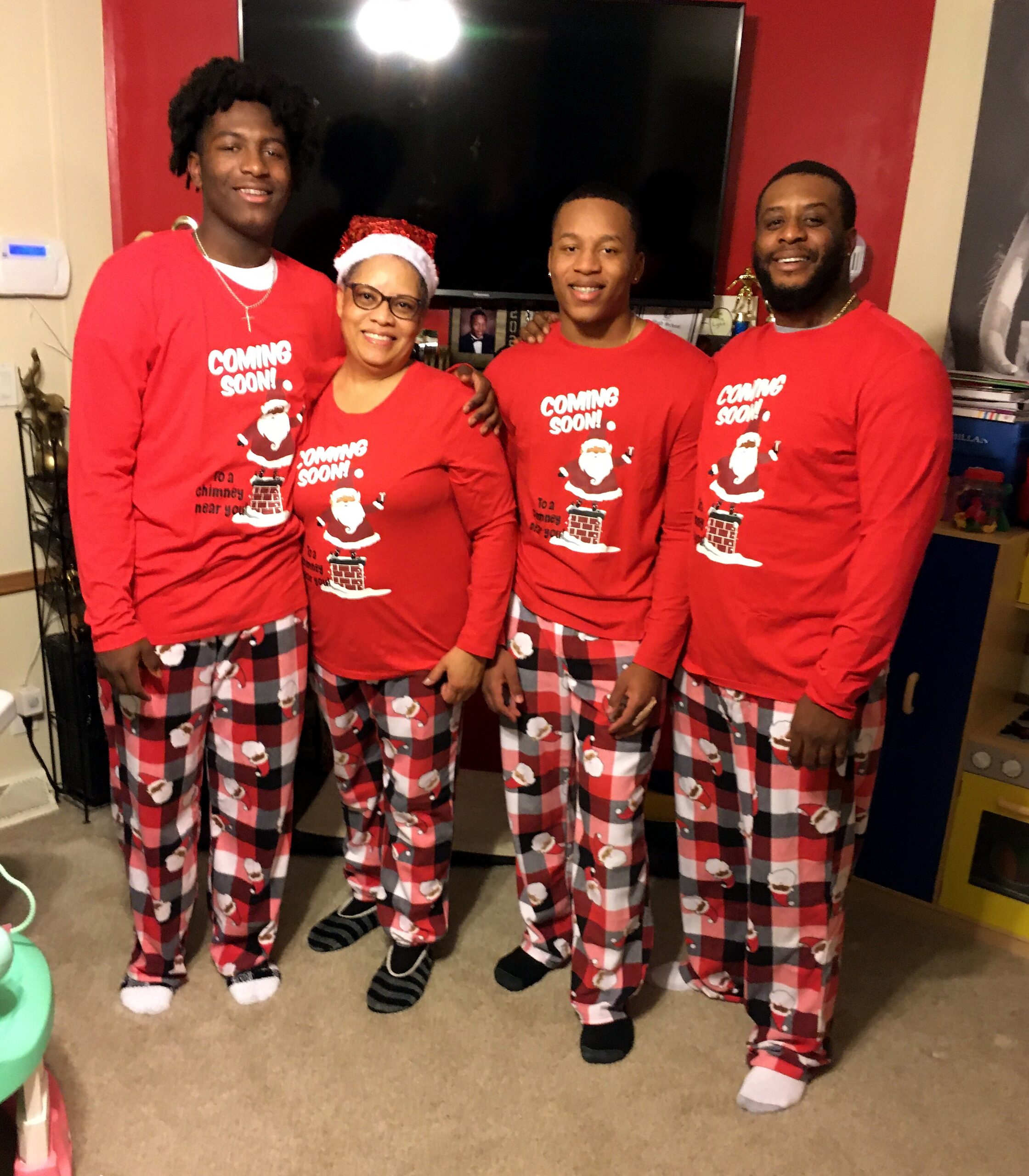 Four people in matching red Christmas pajamas with Santa prints stand together in a living room, smiling at the camera.
