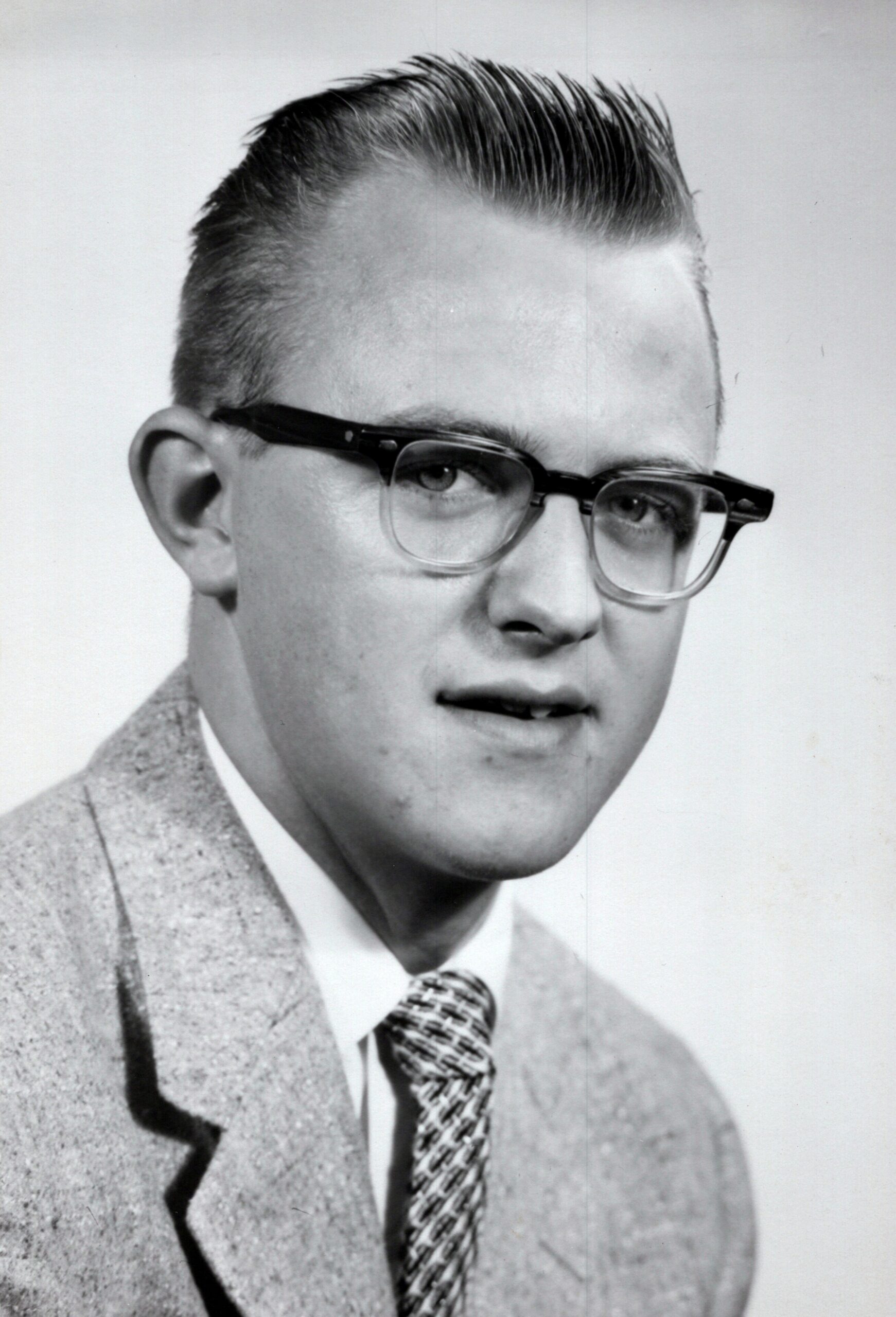 Black-and-white portrait of a young man with slicked-back hair, thick glasses, a light suit, and a patterned tie.