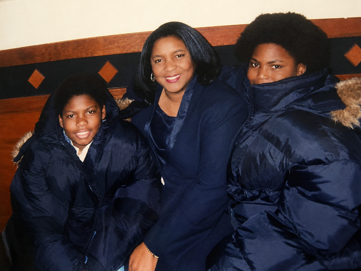 Woman seated between two children, all wearing navy puffer jackets with fur-trimmed hoods, posing for a photo indoors.