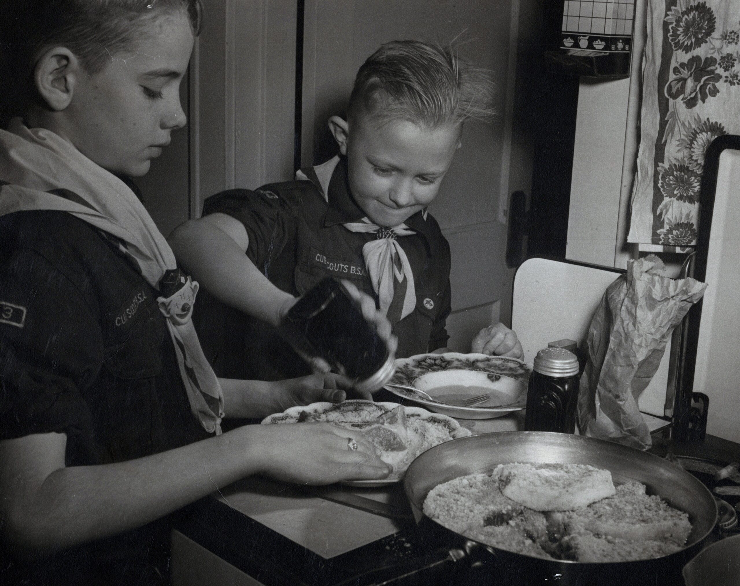 Two young Cub Scouts in uniforms cut and share cake at a kitchen table.