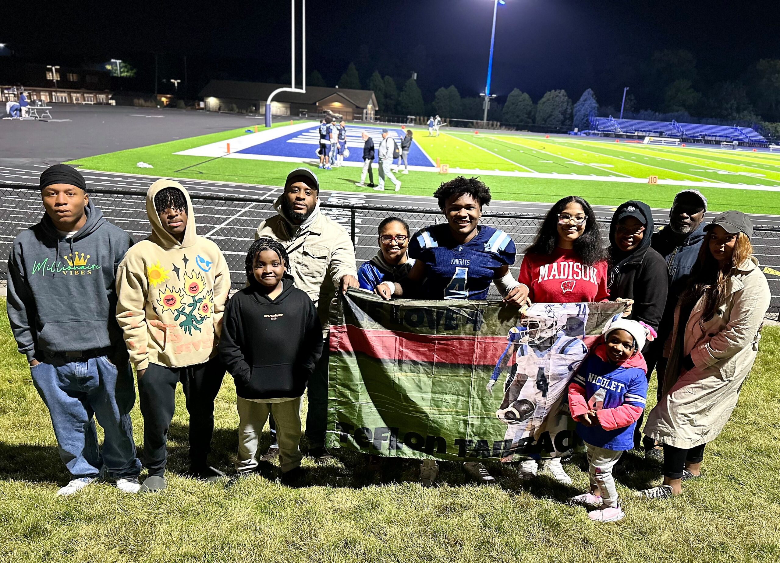 Group of diverse young people posing for a photo in front of a lit football field at night, holding a banner.