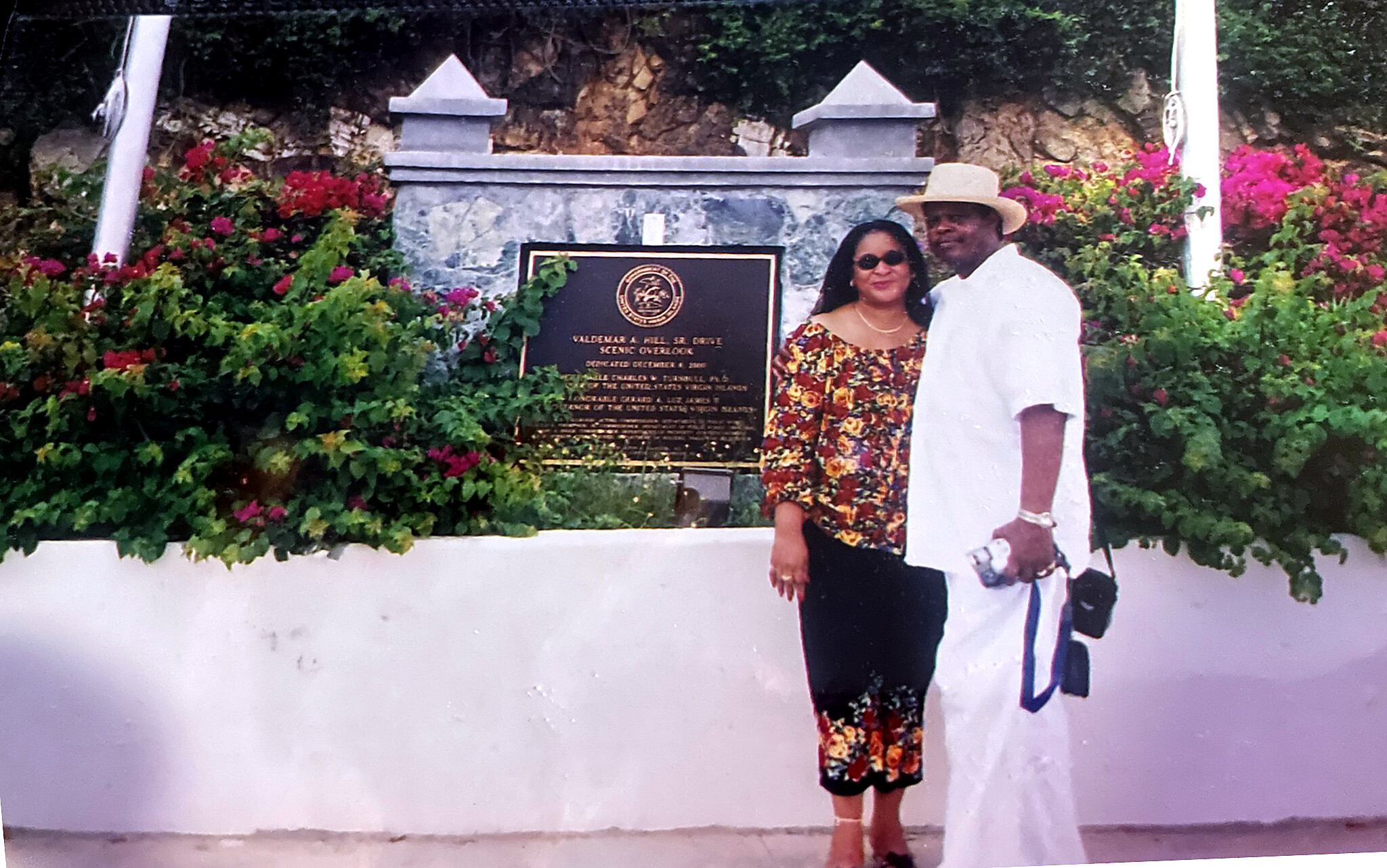 Couple posing by a memorial plaque surrounded by colorful flowers, outdoors, wearing smart casual clothes and hats.