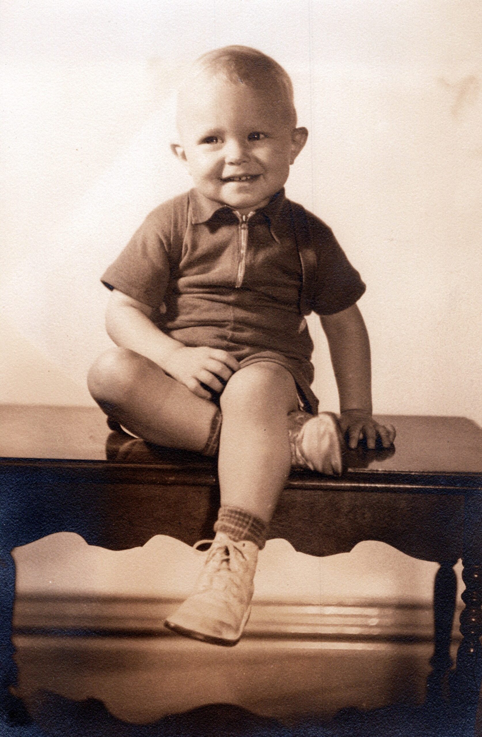 Smiling toddler sitting on a dark wooden bench, wearing a polo shirt and sneakers in a vintage sepia photo.