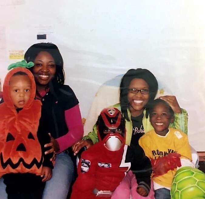 Family posing in Halloween costumes: two adults with two kids, one child in a pumpkin costume and another in a red masked outfit, all smiling.