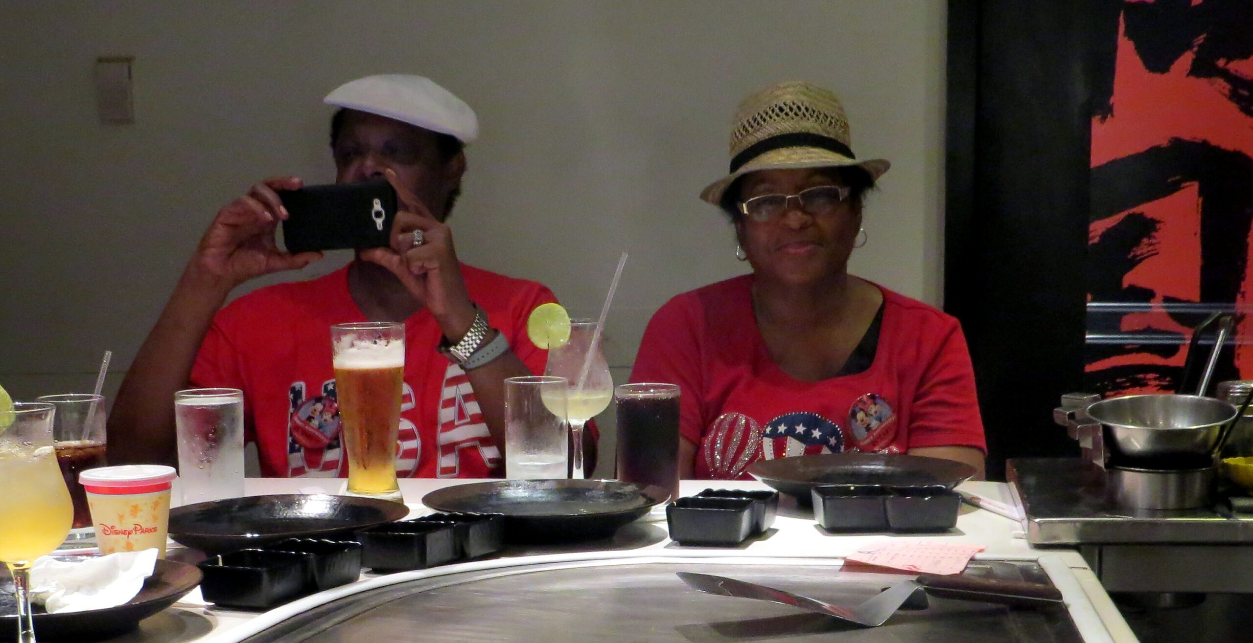 Two women sit at a dining table, one taking a photo with a smartphone while the other wears a straw hat and smiles, both in red shirts.