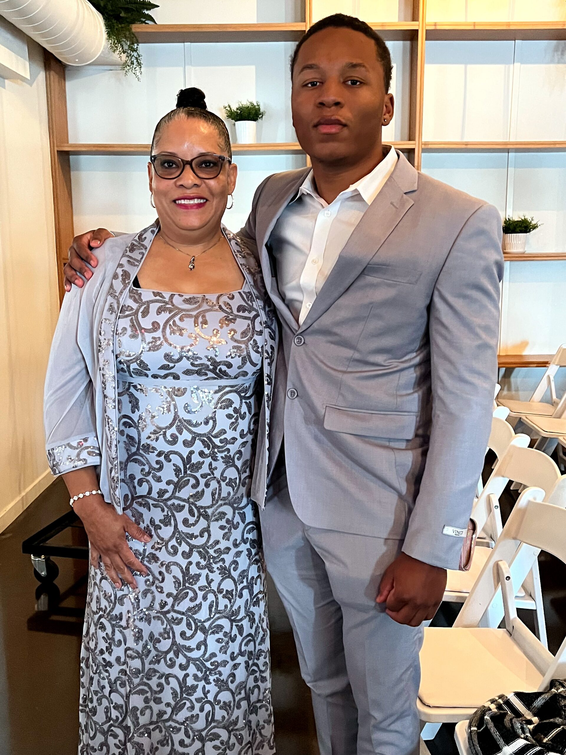 Two people posing together indoors: a smiling woman in a silver patterned dress and a man in a light gray suit, arm around each other.