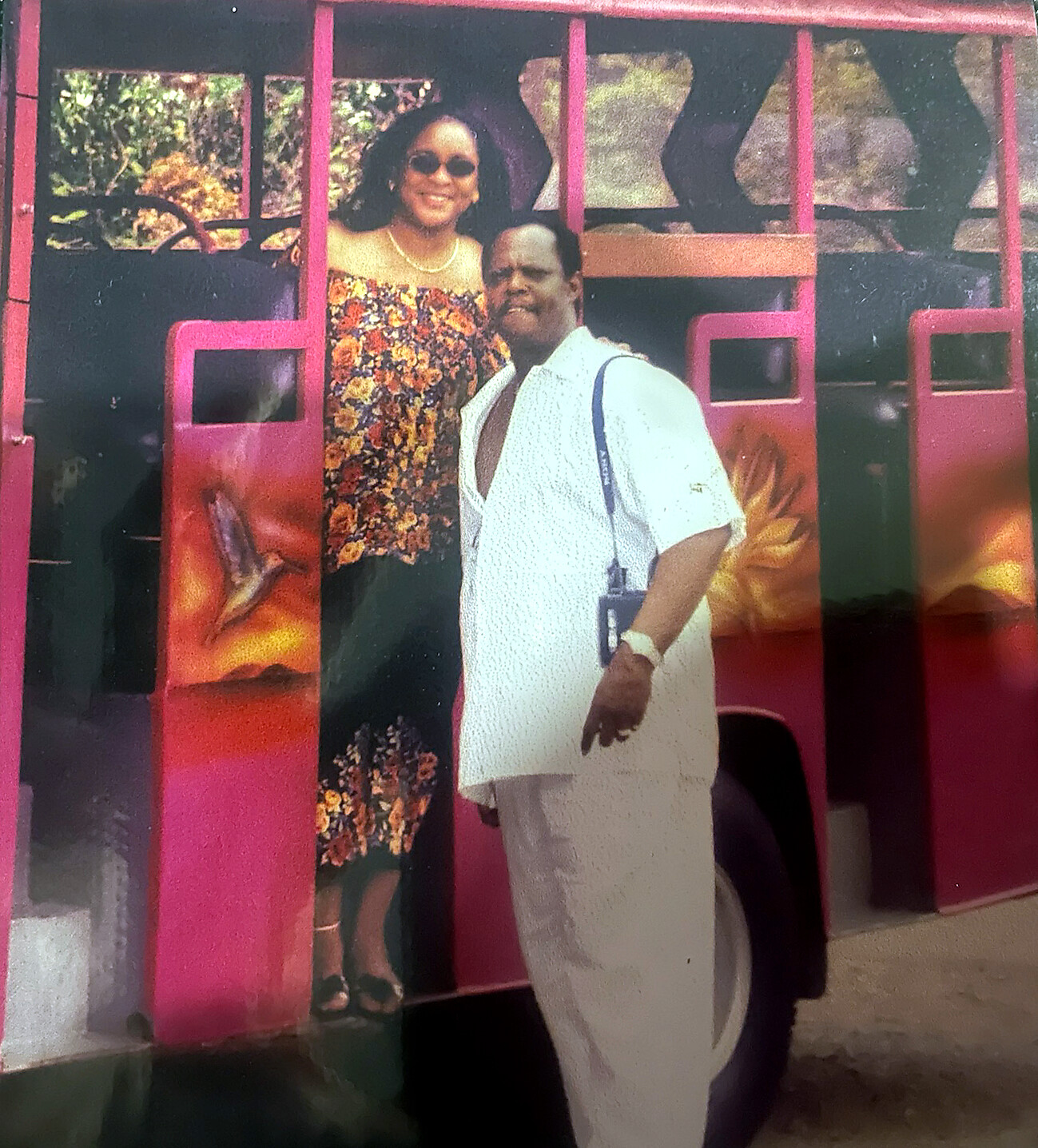 Couple posing in front of a bright pink bus; woman in floral off-shoulder dress and sunglasses, man in a white shirt and light pants.