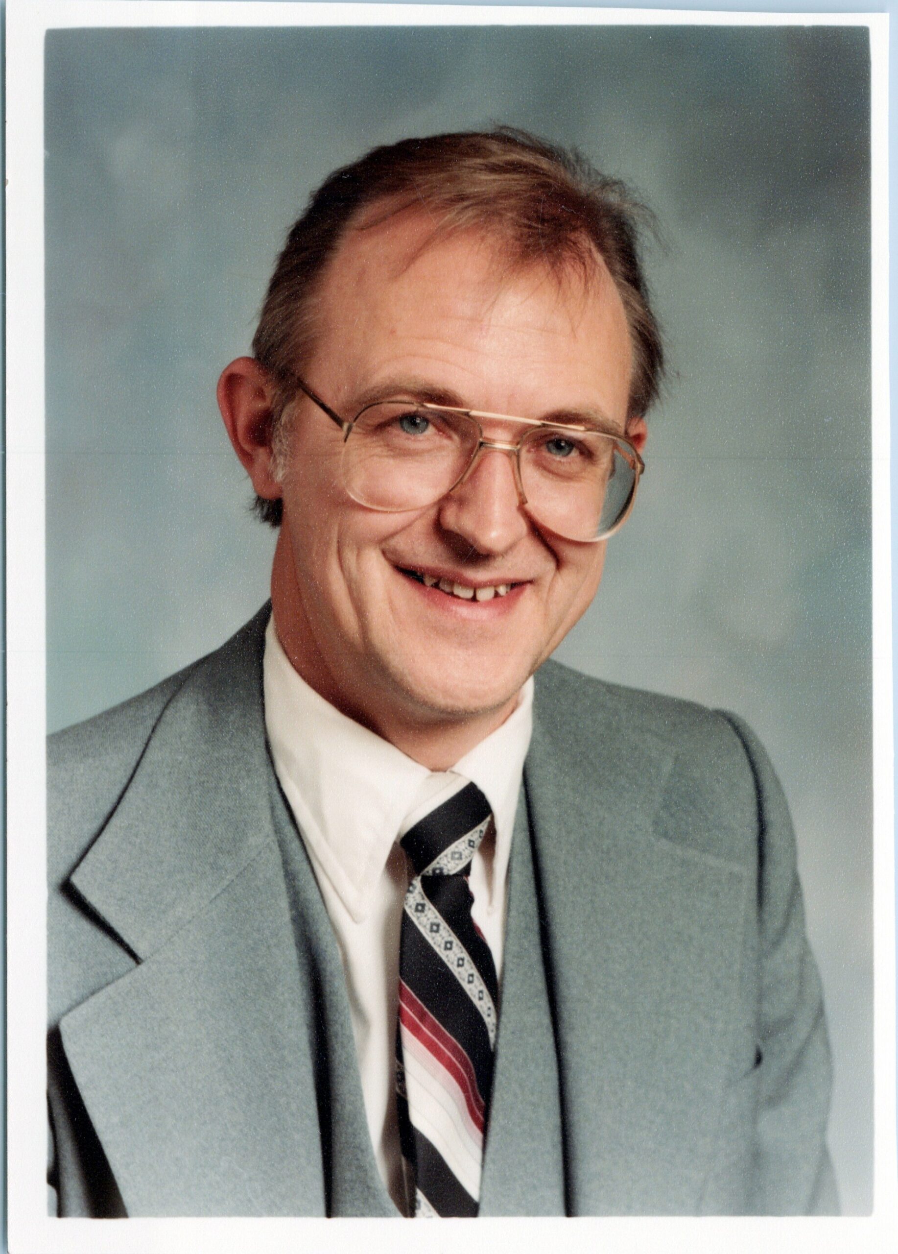 Smiling man in a light gray suit, white shirt, and striped tie, wearing glasses, looking at the camera against a soft gray background.