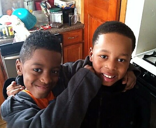Two boys smiling and hugging in a home kitchen, side by side with arms around each other.