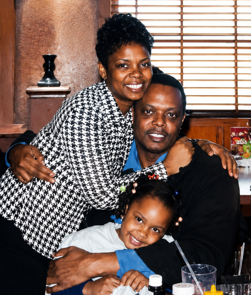 Smiling couple hugs their young daughter at a dining table in a cozy restaurant setting, all three smiling at the camera.