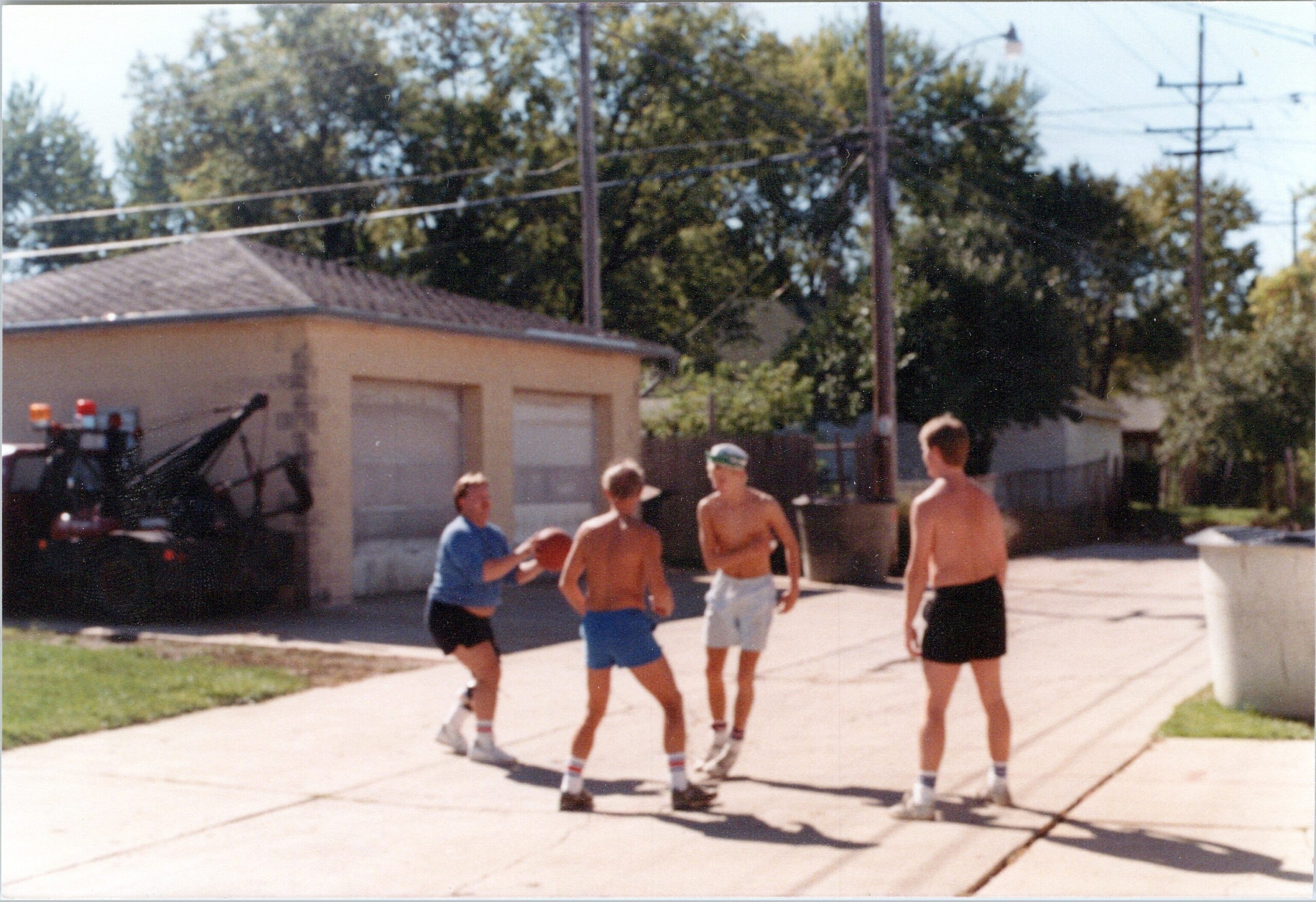 Group of shirtless boys in swim trunks and socks standing on a sunny suburban driveway; a motorcycle with lights is parked to the left, power lines above.