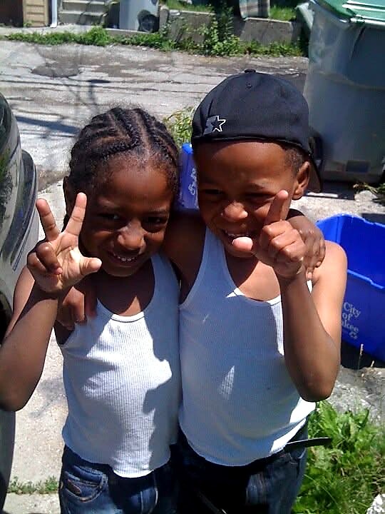 Two kids in white sleeveless shirts smile and show peace signs while standing close together in an outdoor alley.