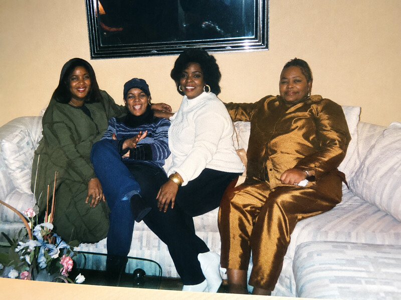 Four women sitting closely on a light-colored sofa, smiling for the camera in a living room setting. From left to right: a woman in a green coat, a woman in a striped blue sweater and cap, a woman in a white turtleneck, and a woman in a brown satin outfit.