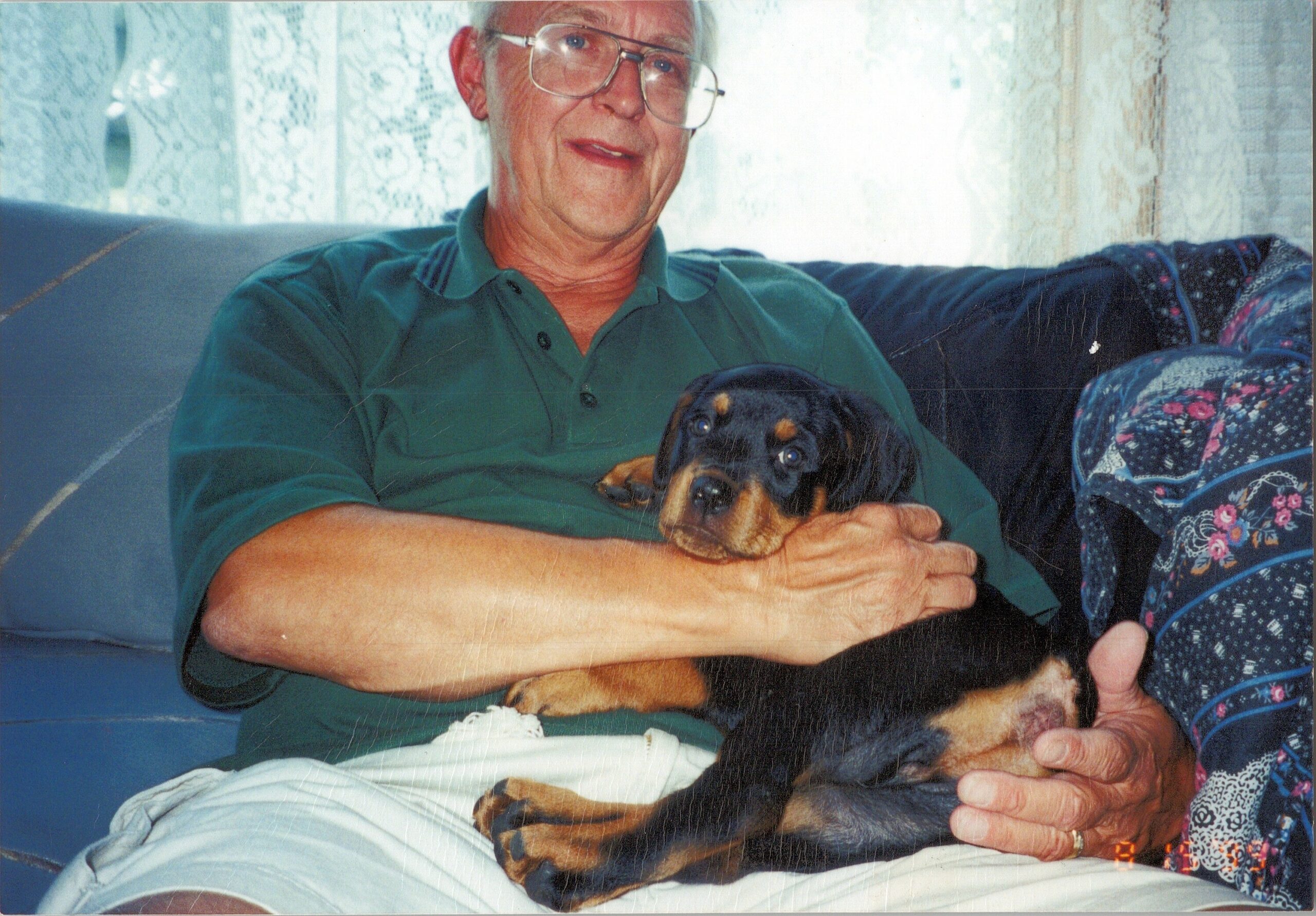 Older man in a green shirt sits on a couch, holding a small black and tan dachshund puppy.