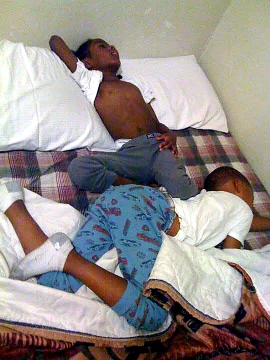 Two young children sleeping on a bed; one shirtless with an arm behind his head, the other in blue patterned pajamas.