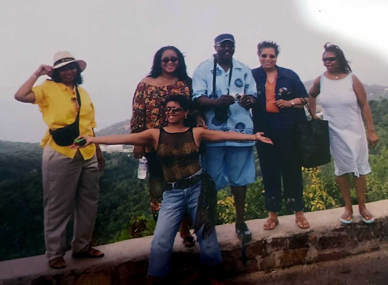 Six friends pose together on a stone ledge with a forested hillside behind them, one person in front with arms outstretched.