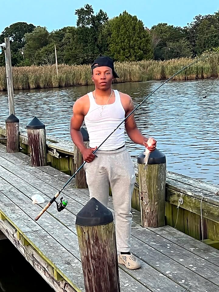Young man standing on a wooden dock by a river, holding a fishing rod with a fish in his hand.