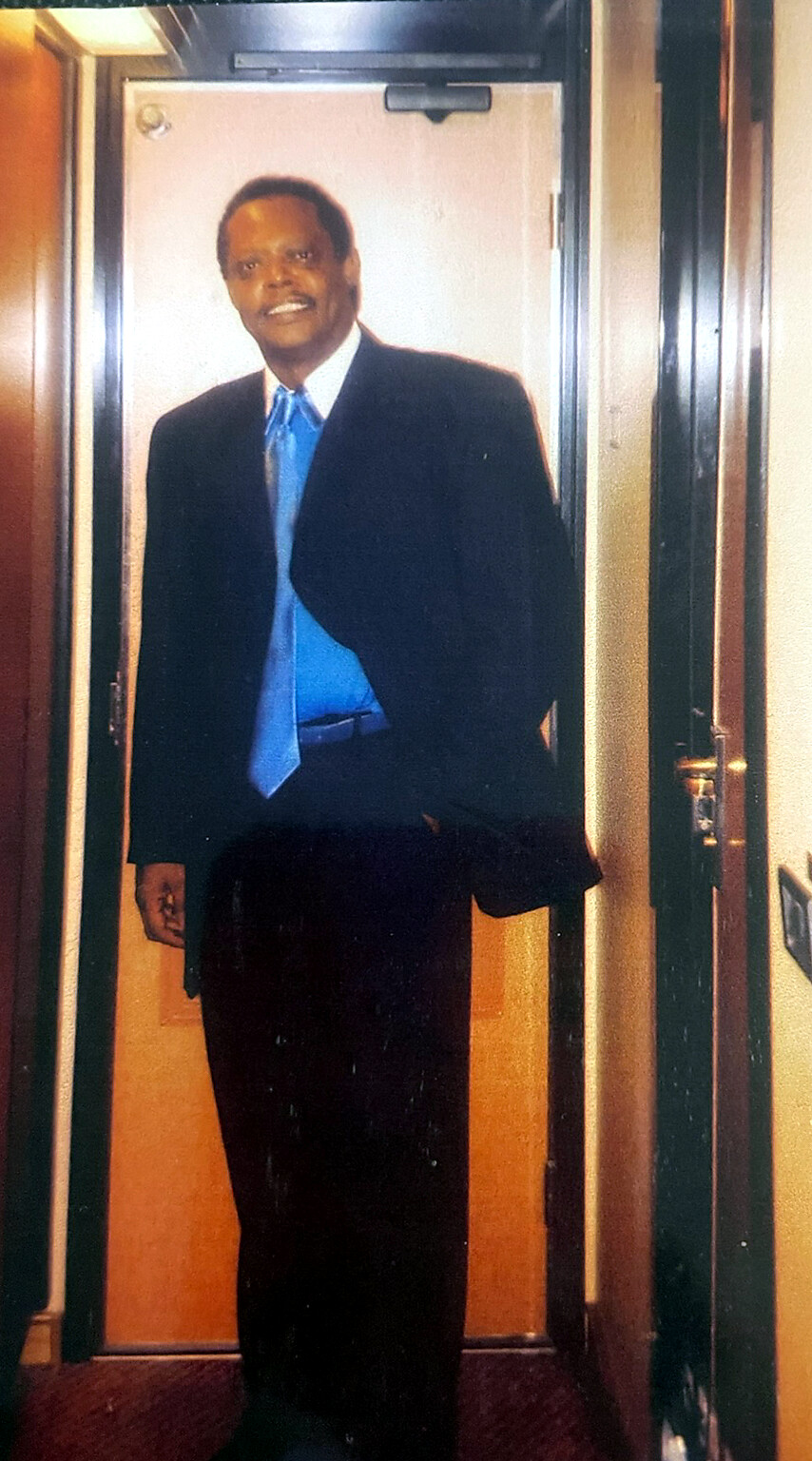 Man in a dark suit and blue tie standing in a hallway near a closed door, smiling.