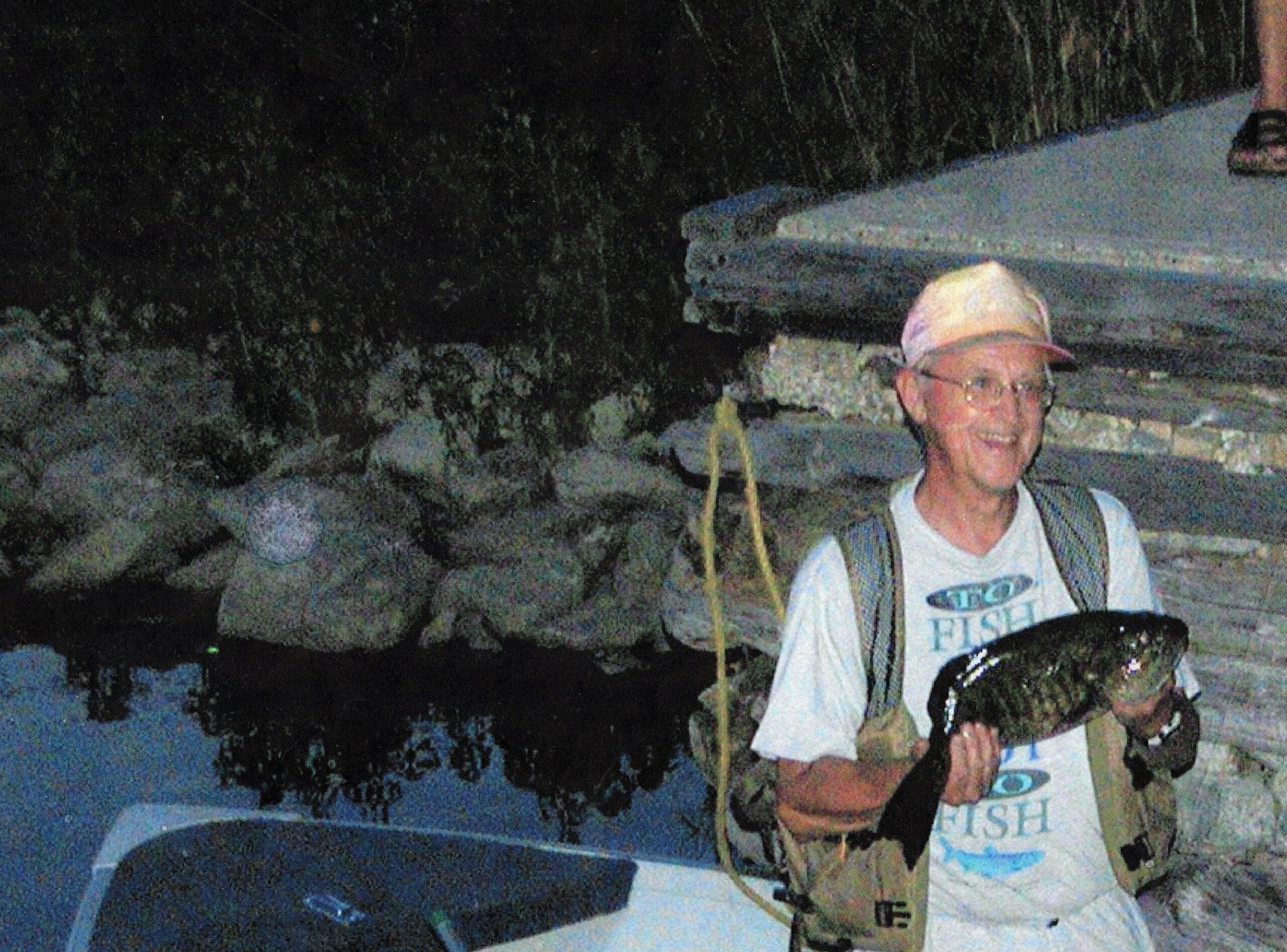 Man wearing a hat and life vest on a small boat at night, holding a rope, with dark water and trees in the background.
