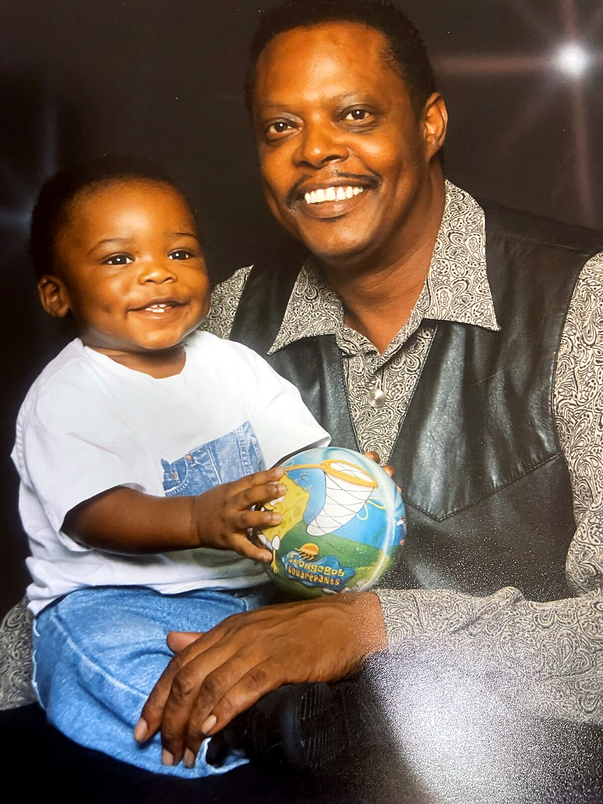 Smiling man with a child on his lap, both looking at the camera, the child holding a colorful ball.