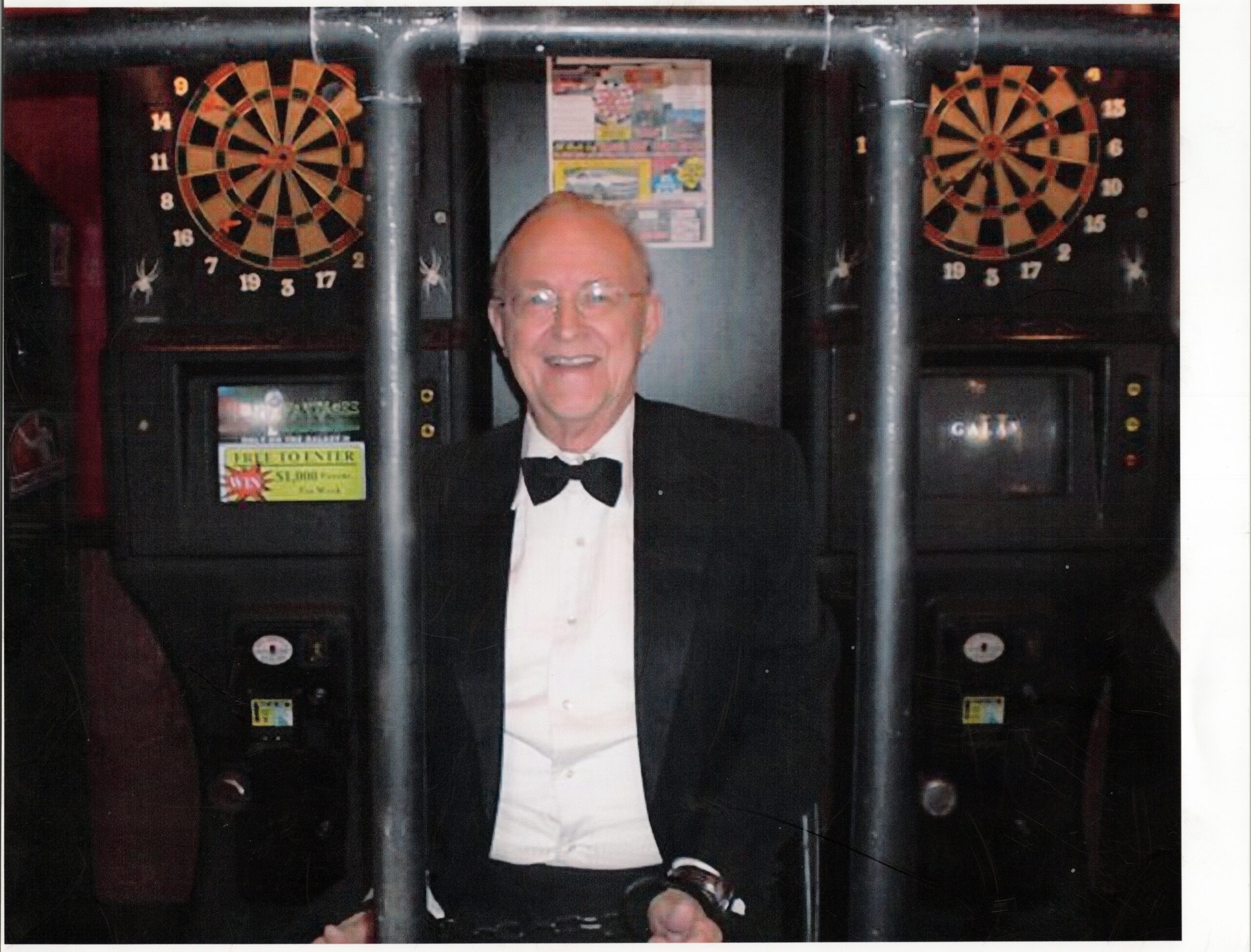Smiling man in a tuxedo standing between two dartboard arcade machines in a dimly lit venue, facing the camera.