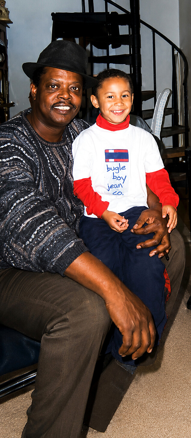 Smiling man in a black hat sits with a young boy in a white shirt and red sleeves on a couch, a spiral staircase in the background.