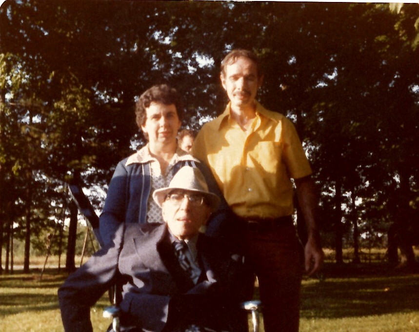 Three people pose in a sunny park: an elderly man in a suit and hat seated in a wheelchair, with a woman and a man standing behind him.
