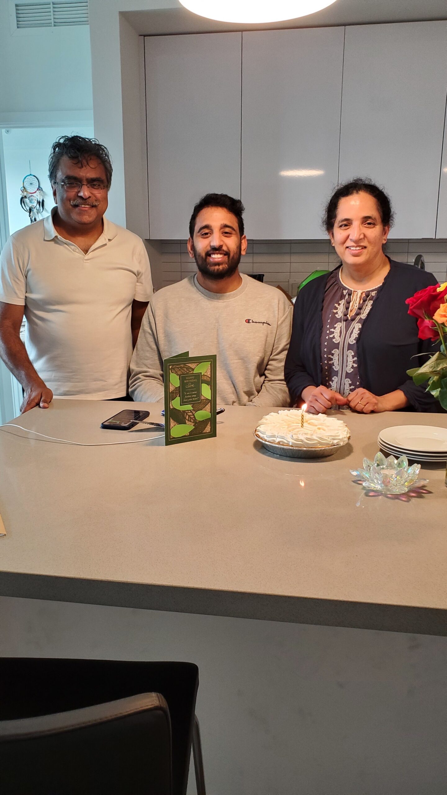 Three people stand behind a birthday cake with a lit candle on a kitchen island, smiling at the camera. A greeting card and a bouquet are nearby.