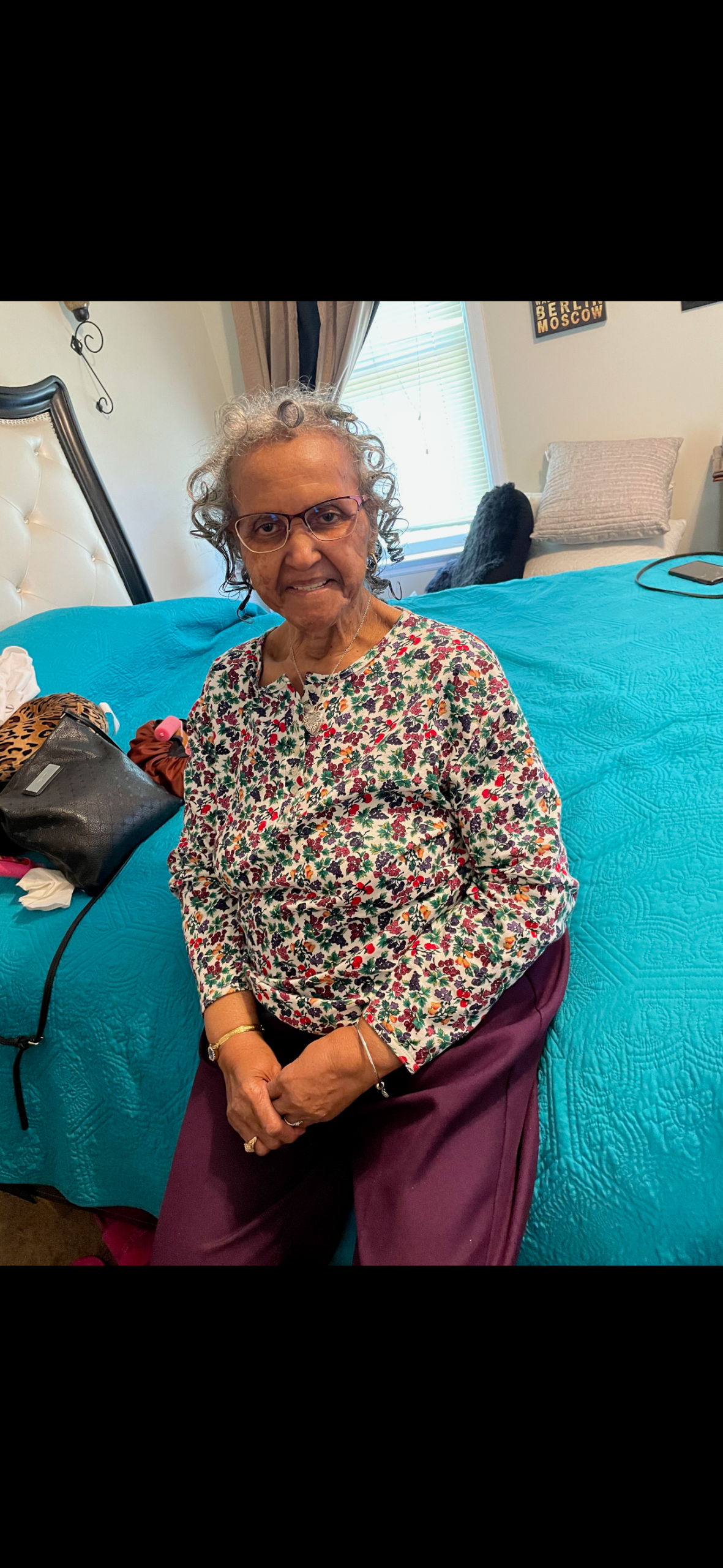 Older woman with short gray curly hair and glasses sits on a bed in a bright bedroom, wearing a colorful floral blouse and purple pants.