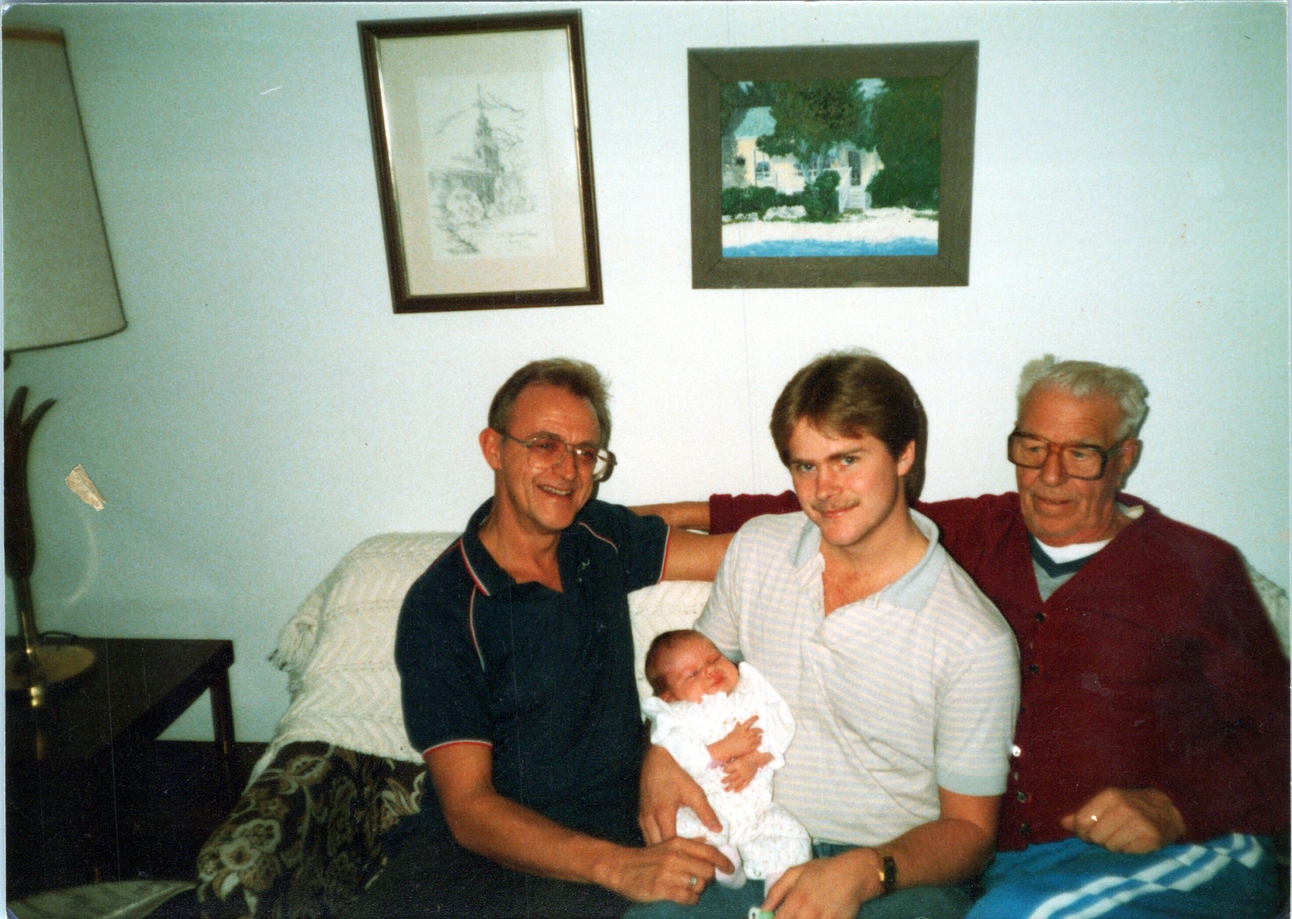 Three men sit on a couch in a living room holding a newborn baby wrapped in a blanket between them, smiling at the camera.