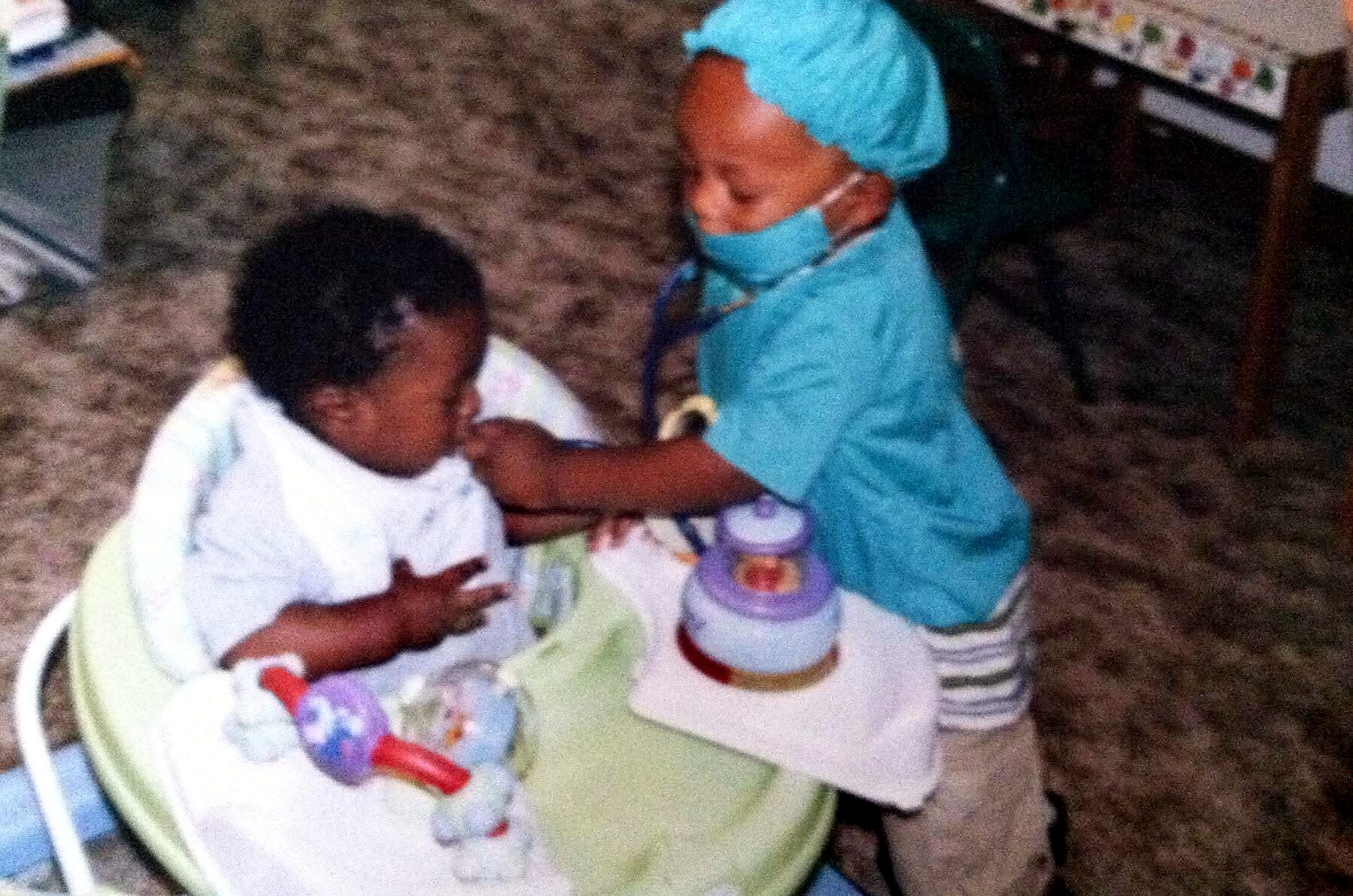 Nurse in blue scrubs and cap leans over a child in a high chair, mask on, with a stethoscope nearby during care at a table.