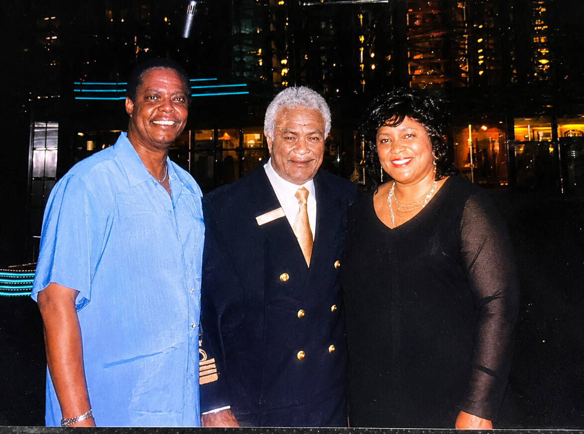 Three adults posing together outdoors at night, smiling, with a city-lit backdrop: man in blue shirt left, man in navy suit center, woman in black dress right.