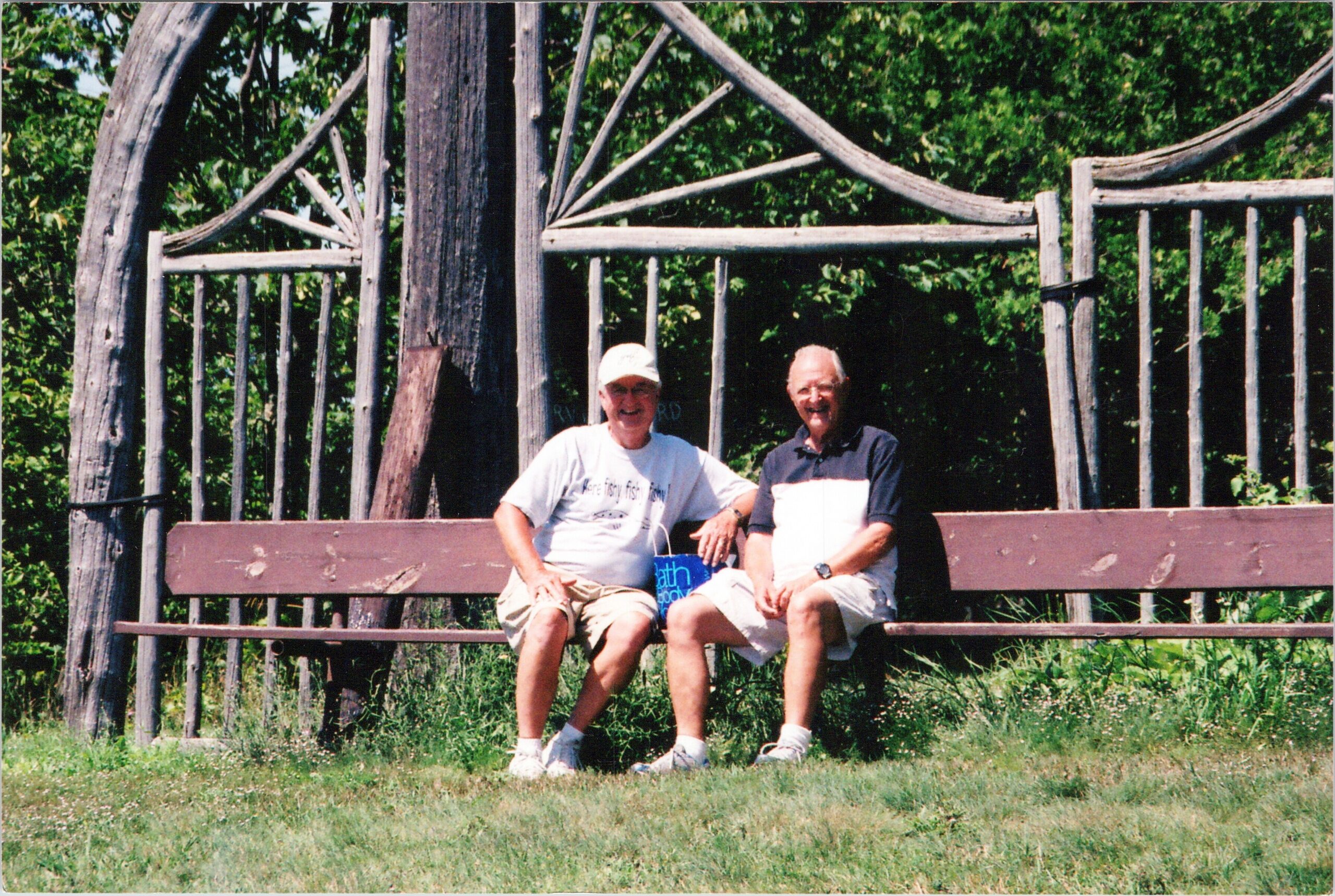 Two older men sit on a weathered wooden fence outdoors, smiling with green trees in the background.