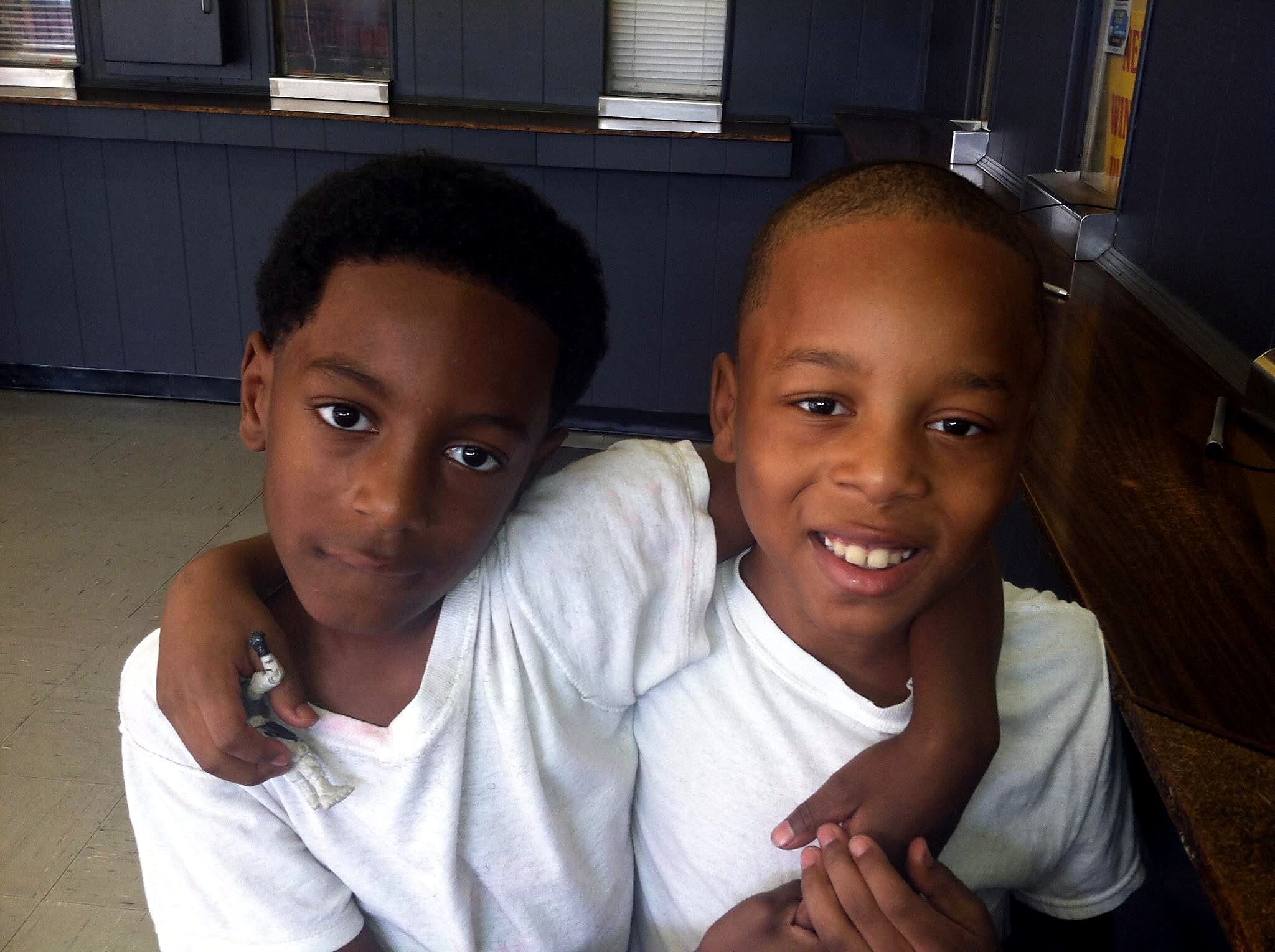 Two smiling boys pose closely together, their arms around each other, indoors in front of blue walls.