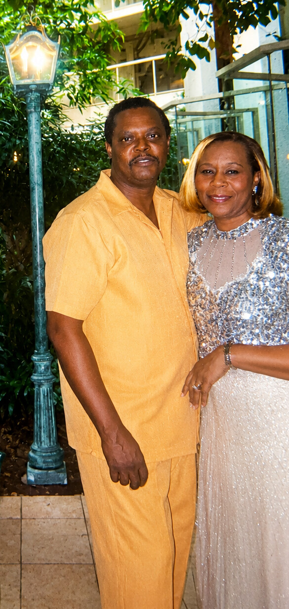 Couple posing together outdoors at night, man in a yellow shirt and pants and woman in a sparkling silver dress, smiling at the camera near a lamp post.