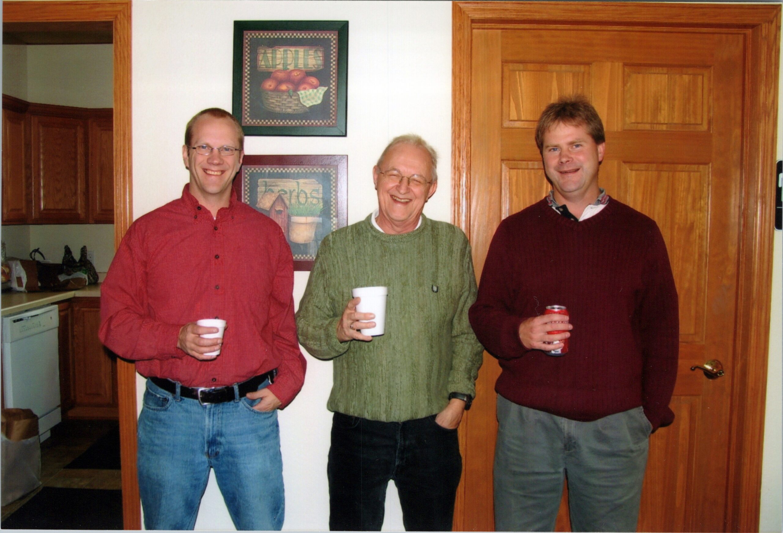 Three men stand side by side in a home kitchen, each holding a cup and smiling at the camera.