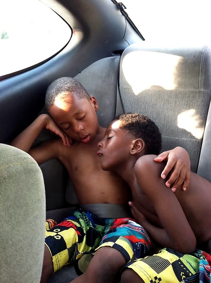 Two shirtless boys asleep in the back seat of a car, cuddled together with arms around each other.