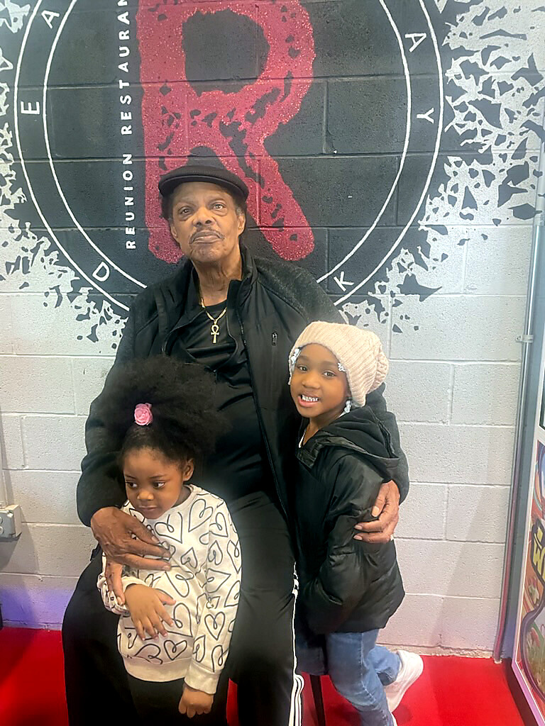 An older man wearing a cap sits with two young children, one on each side, posing for a photo in front of a wall with a circular logo behind them.
