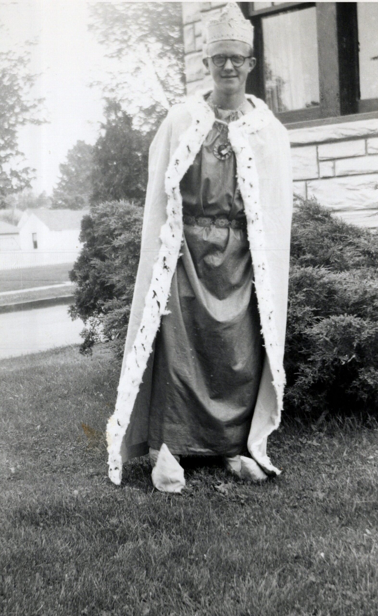 Person in a king costume with crown and long cape, smiling on a lawn in front of a stone house.