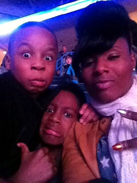 Three people taking a selfie at a crowded venue; two adults and a child make funny faces at the camera.