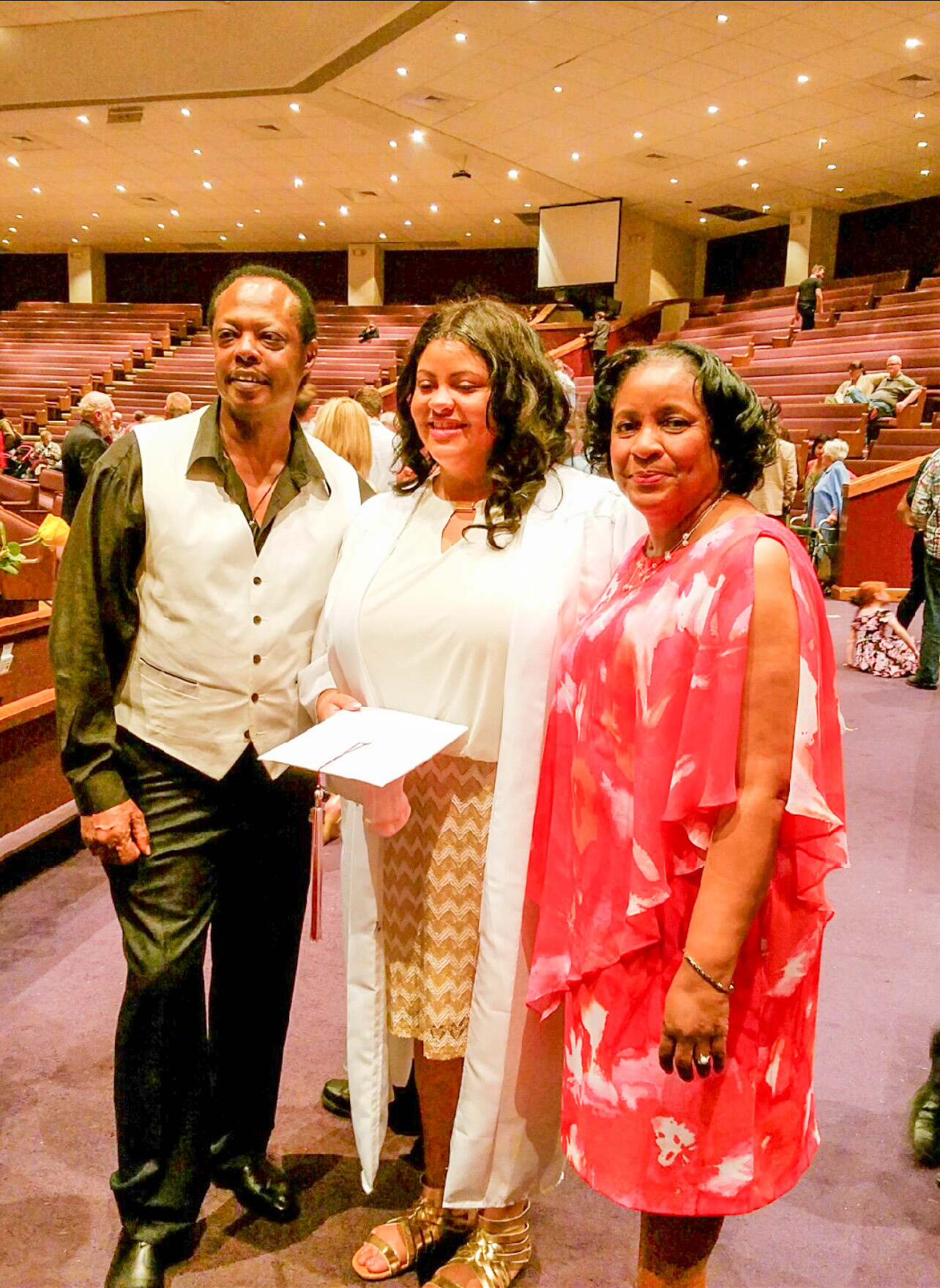 Three adults posing together in a church or auditorium after a ceremony, smiling at the camera.