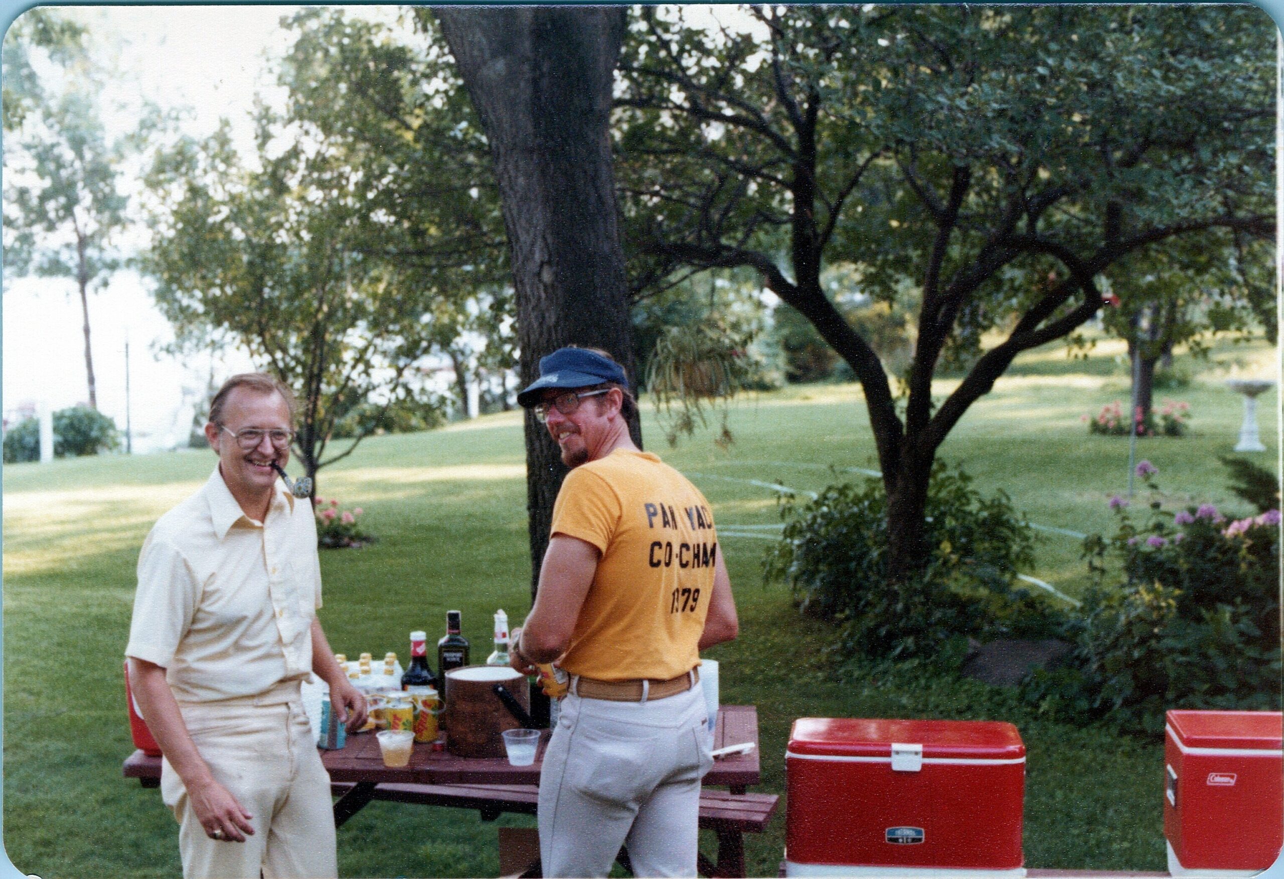 Two men stand by a picnic table with drinks on a sunny lawn, red coolers nearby, trees in the background.